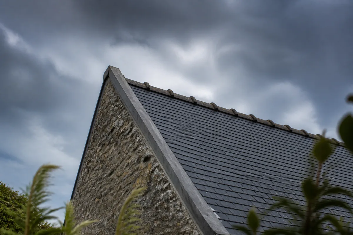 Toiture en ardoise d’une maison en pierre exposée au vent, ciel de littoral chargé